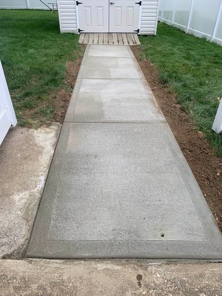 Concrete walkway leading to a white shed, flanked by grass and dirt.