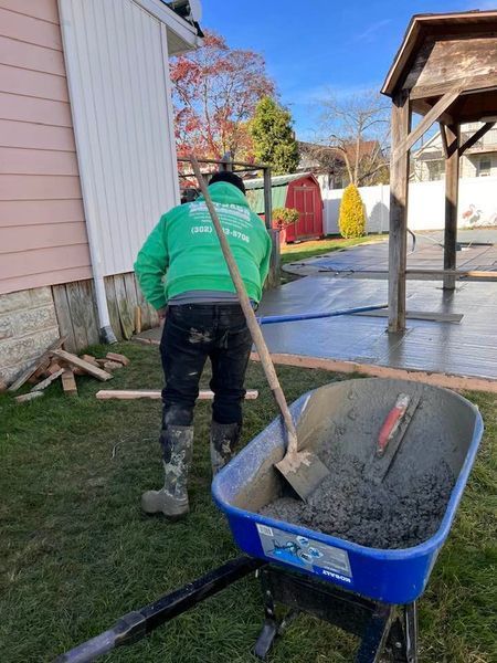 A person in green jacket shovels concrete from a wheelbarrow onto a patio, next to a pink house and gazebo.