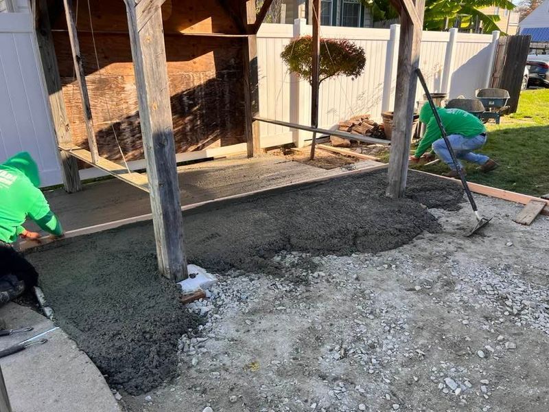 Workers in green shirts pour and smooth wet concrete around a wooden structure in a backyard.