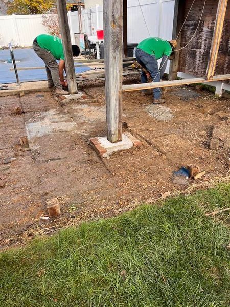 Two workers in green shirts digging in dirt near a metal pole and wooden structure.