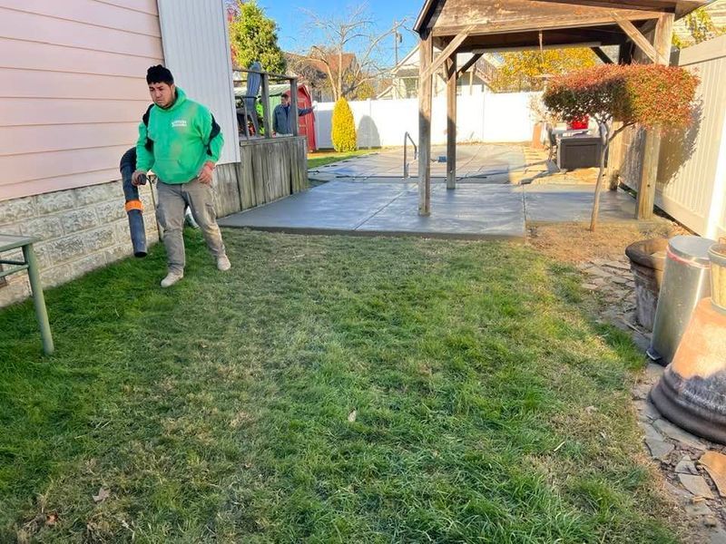 A person in a green sweatshirt walks across a grassy yard toward a concrete patio; another person is near a wall.