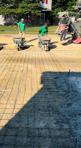 Workers pouring concrete on rebar grid, preparing a paved surface; sunny outdoor setting.