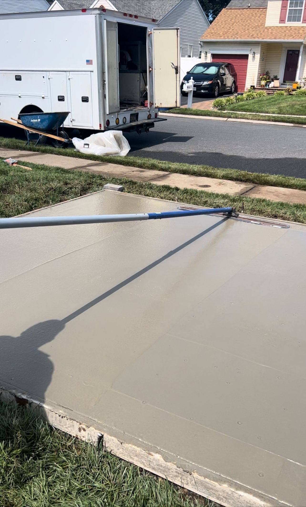 Freshly poured concrete being smoothed with a long tool on a driveway, utility truck in background.