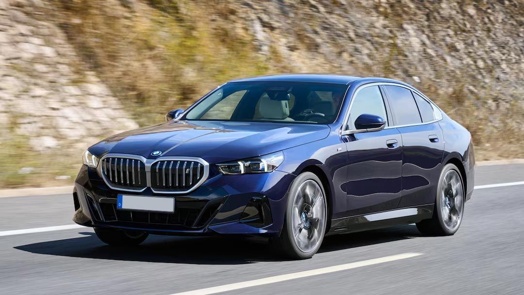 A blue bmw is driving down a road next to a mountain.