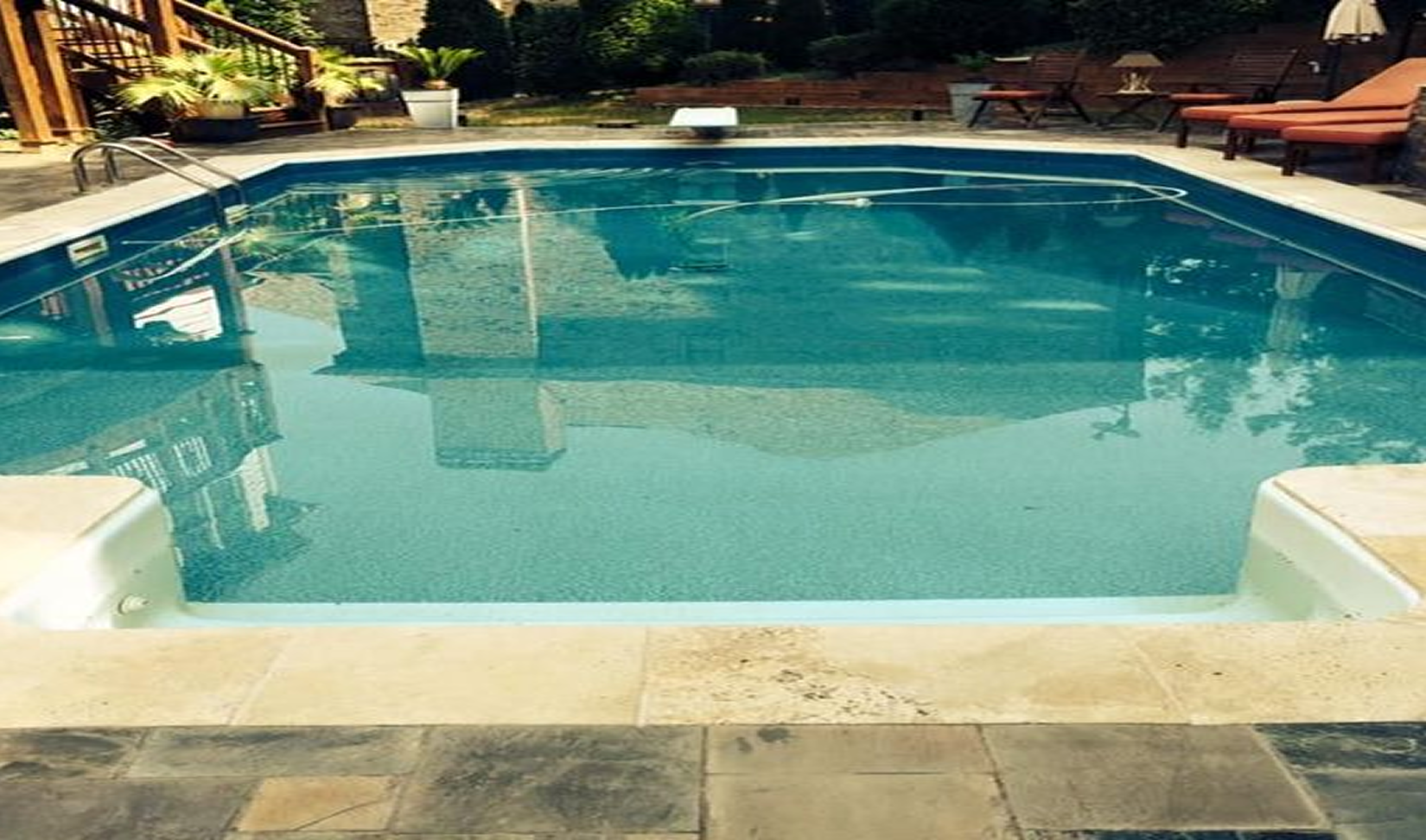Swimming pool surrounded by stone patio, deck, and trees; calm blue water.