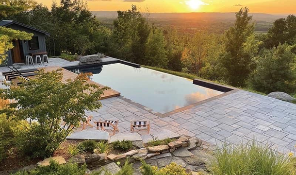Infinity pool overlooking a valley at sunset. Poolside patio with lounge chairs; trees frame the view.