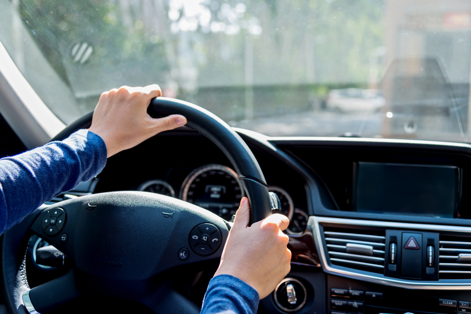 A person's hands gripping the steering wheel of a car while driving on a bright day.