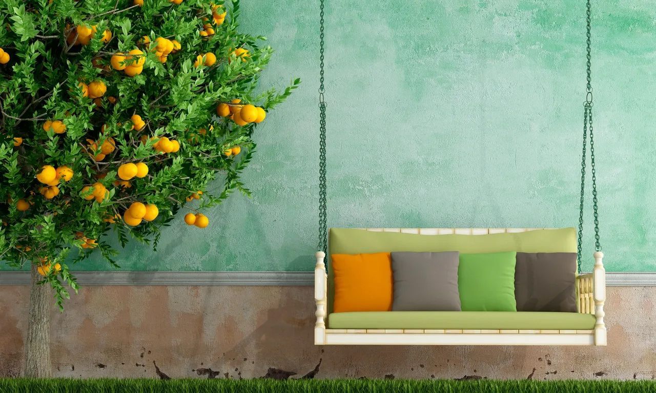 Swing bench with colorful pillows hanging in front of a textured turquoise wall, under a tree laden with yellow fruit.