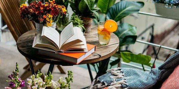 Round wooden table with books, a drink, and flowers on a balcony.