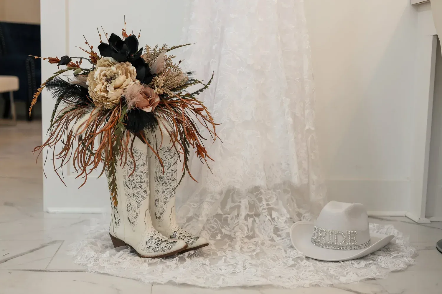 A white lace bridal gown backdrop with white embroidered boots holding a rustic floral bouquet and a beaded cowboy hat.