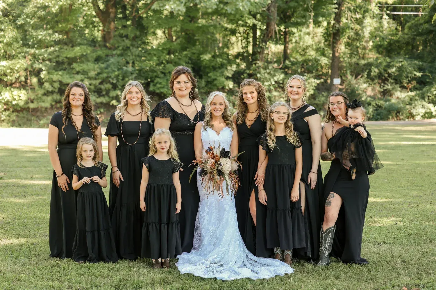 A bride in a white lace gown stands in a grassy field surrounded by ten others wearing black formal dresses.