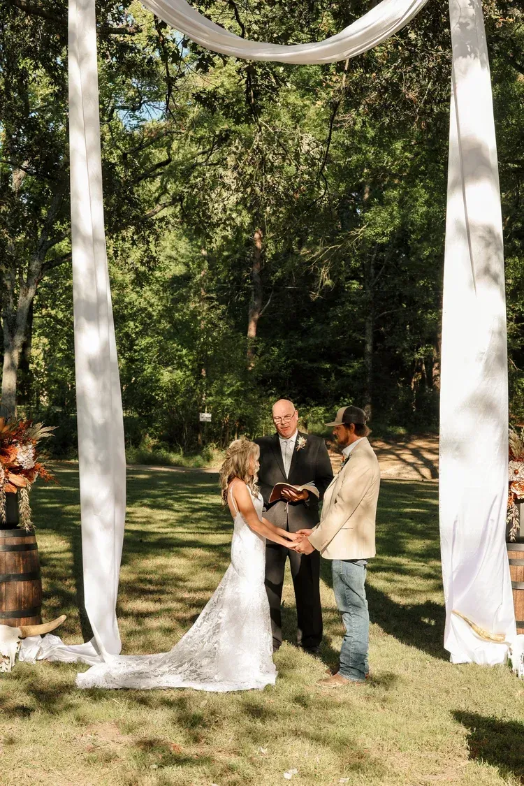 A couple stands together during an outdoor wedding ceremony under a white fabric archway in a wooded area.