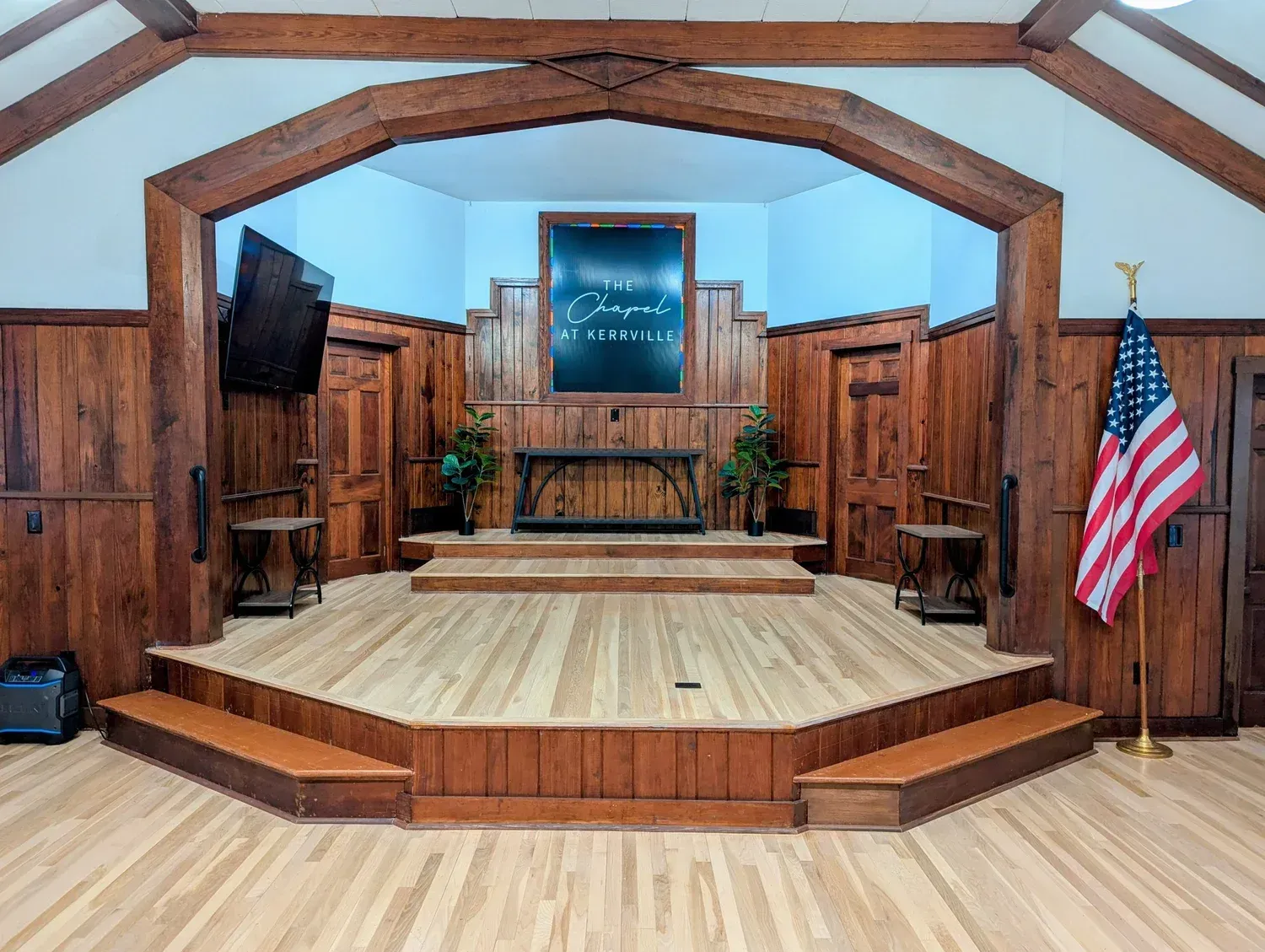 A chapel stage with wood paneling, a raised platform, stairs, a central sign, two small plants, and a US flag.