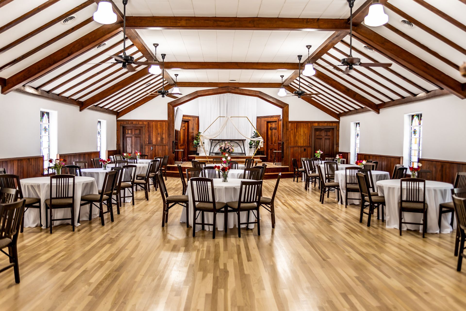 An elegant dining room with round tables, chairs, and stained-glass windows, featuring a screen displaying a photo collage.
