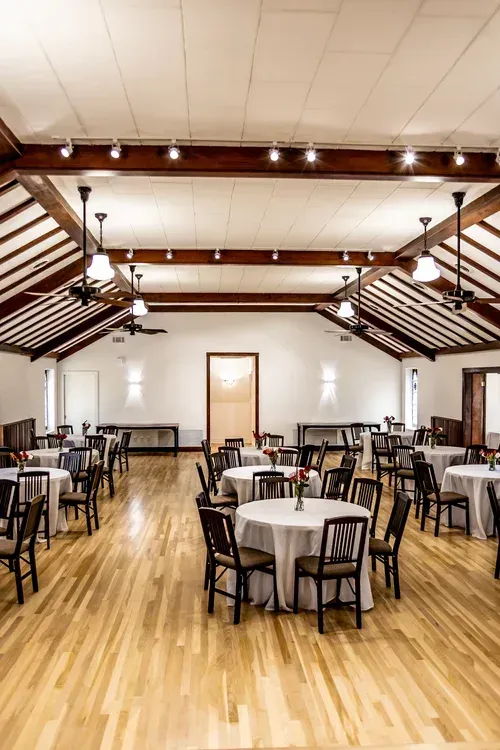 A spacious event hall with high ceilings, exposed wood beams, hardwood floors, and multiple round tables with white cloths.