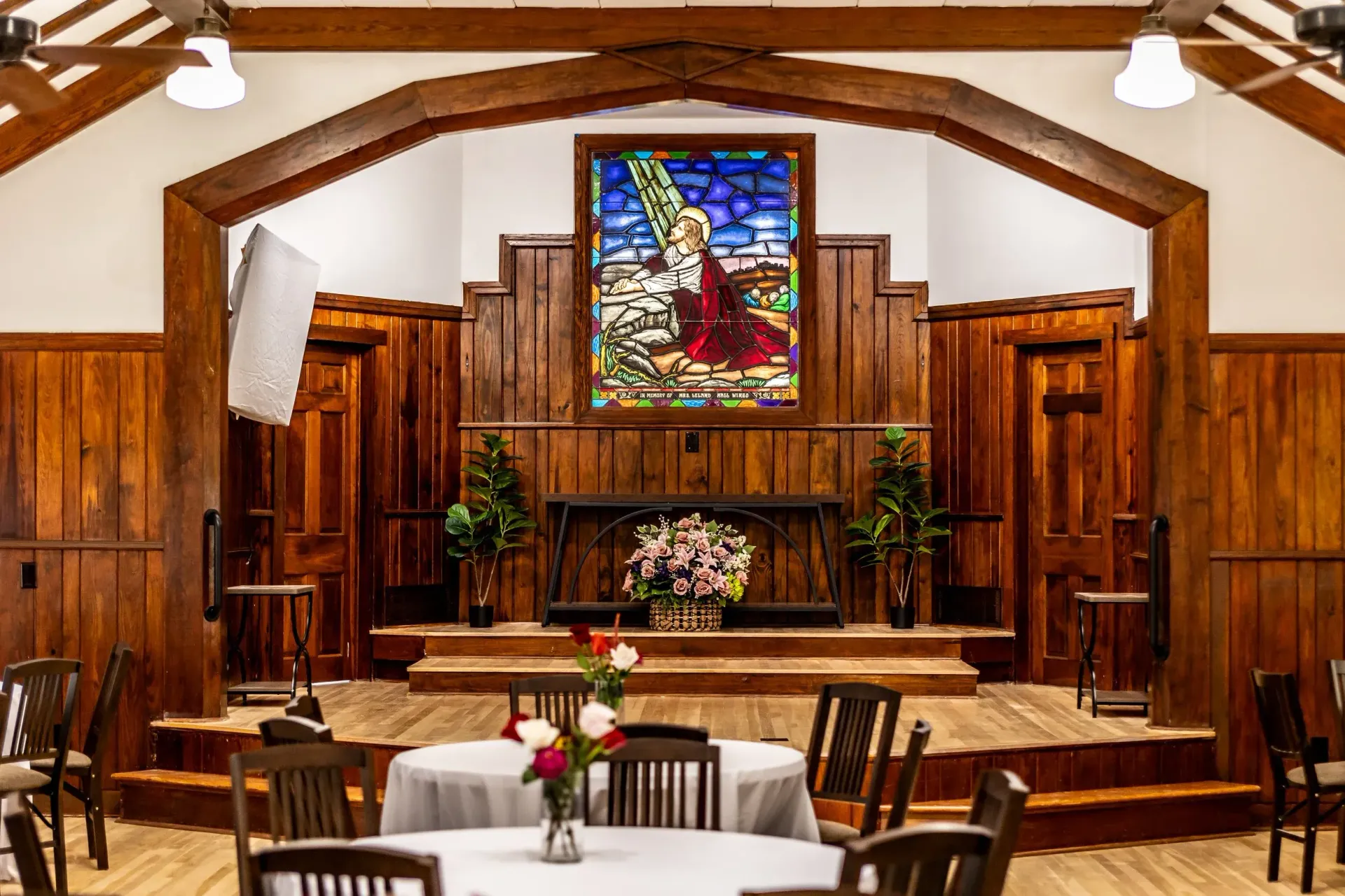 A rustic church interior with wood-paneled walls, a stage, and a stained glass window depicting a figure in a red robe.
