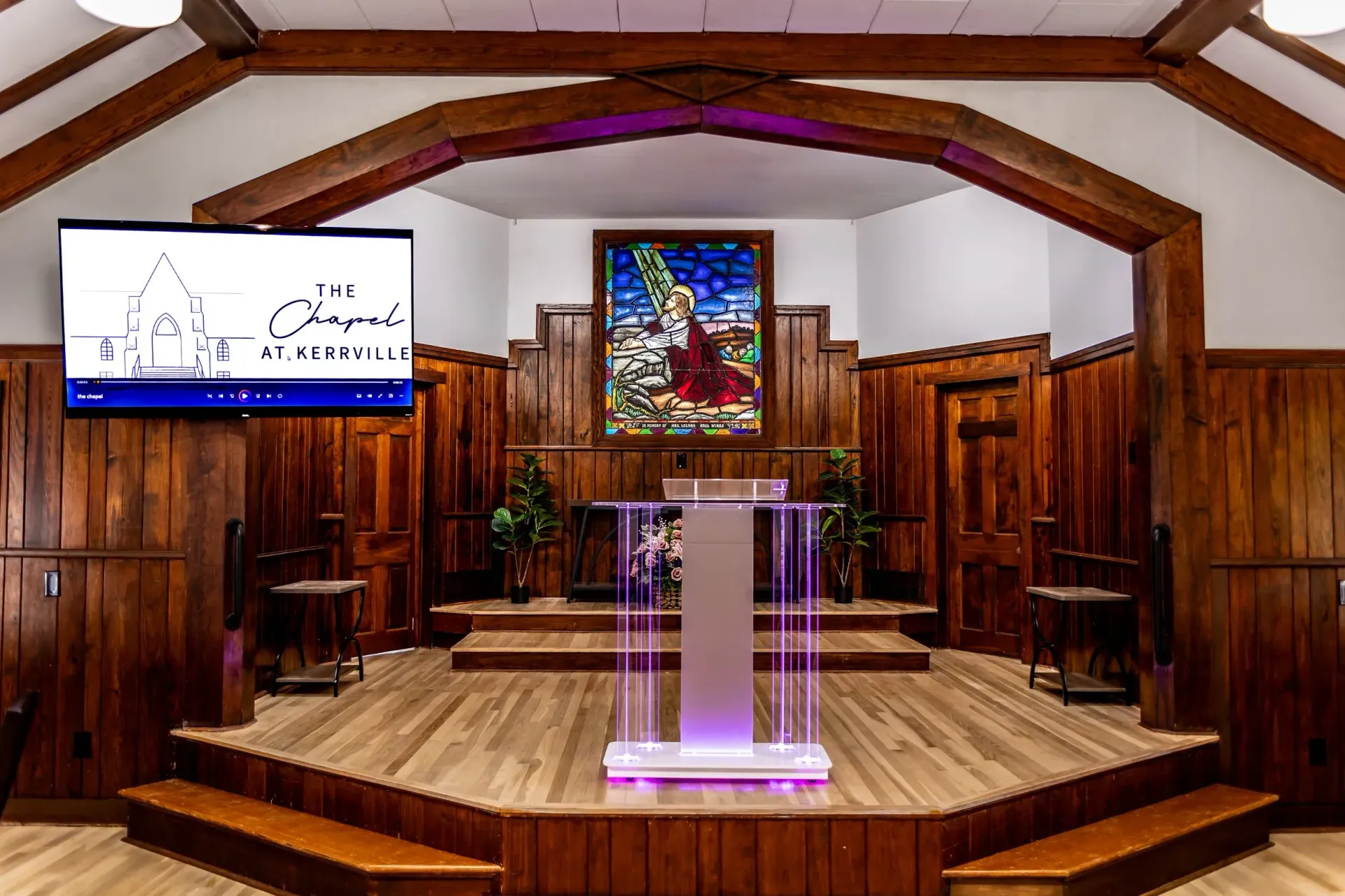A brightly lit church interior featuring a wooden pulpit on a stage, a stained-glass window, and a wall-mounted display.