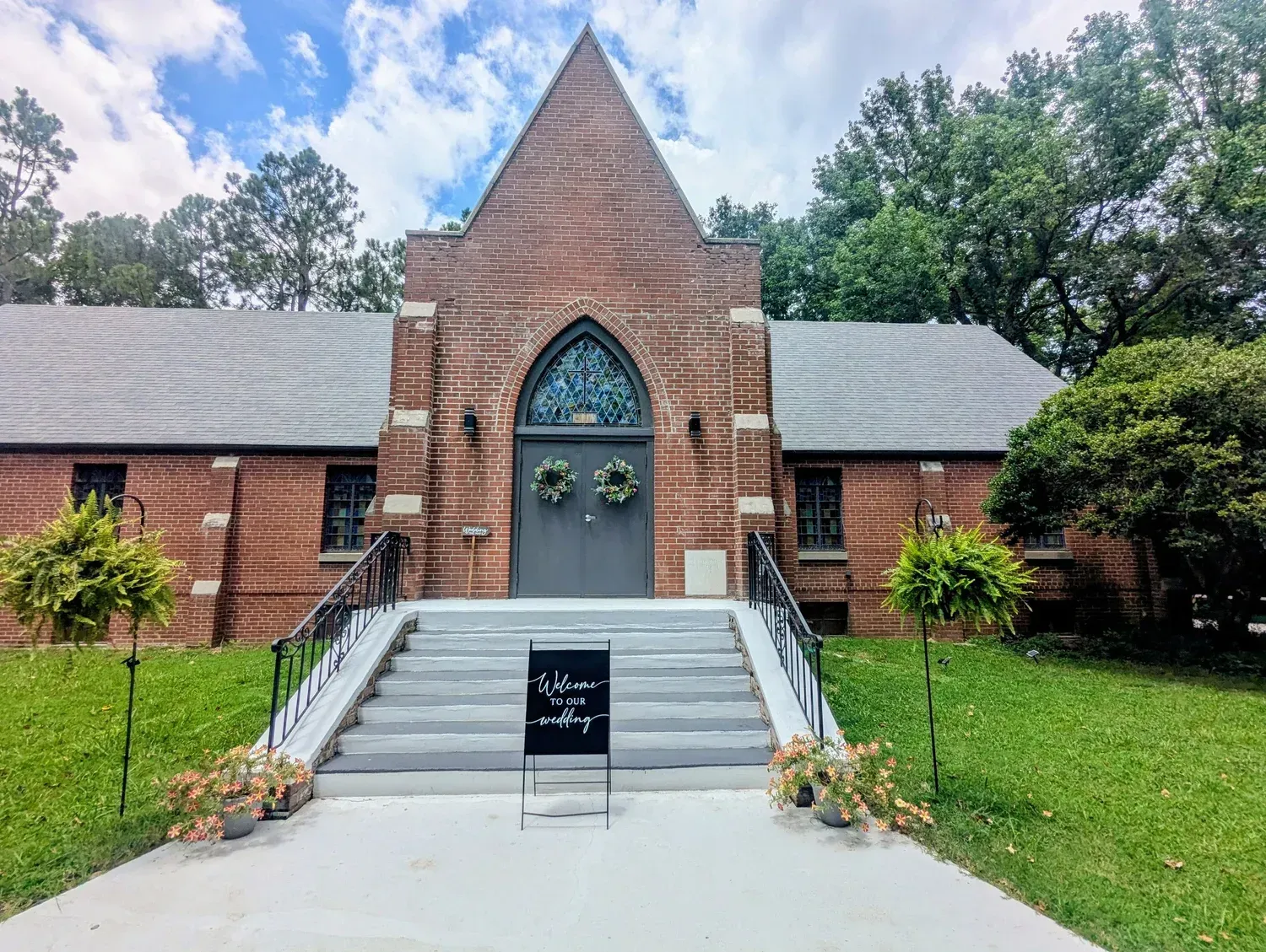 Brick church with a grey roof and front steps, adorned with two wreaths on the grey door and hanging plants on each side.
