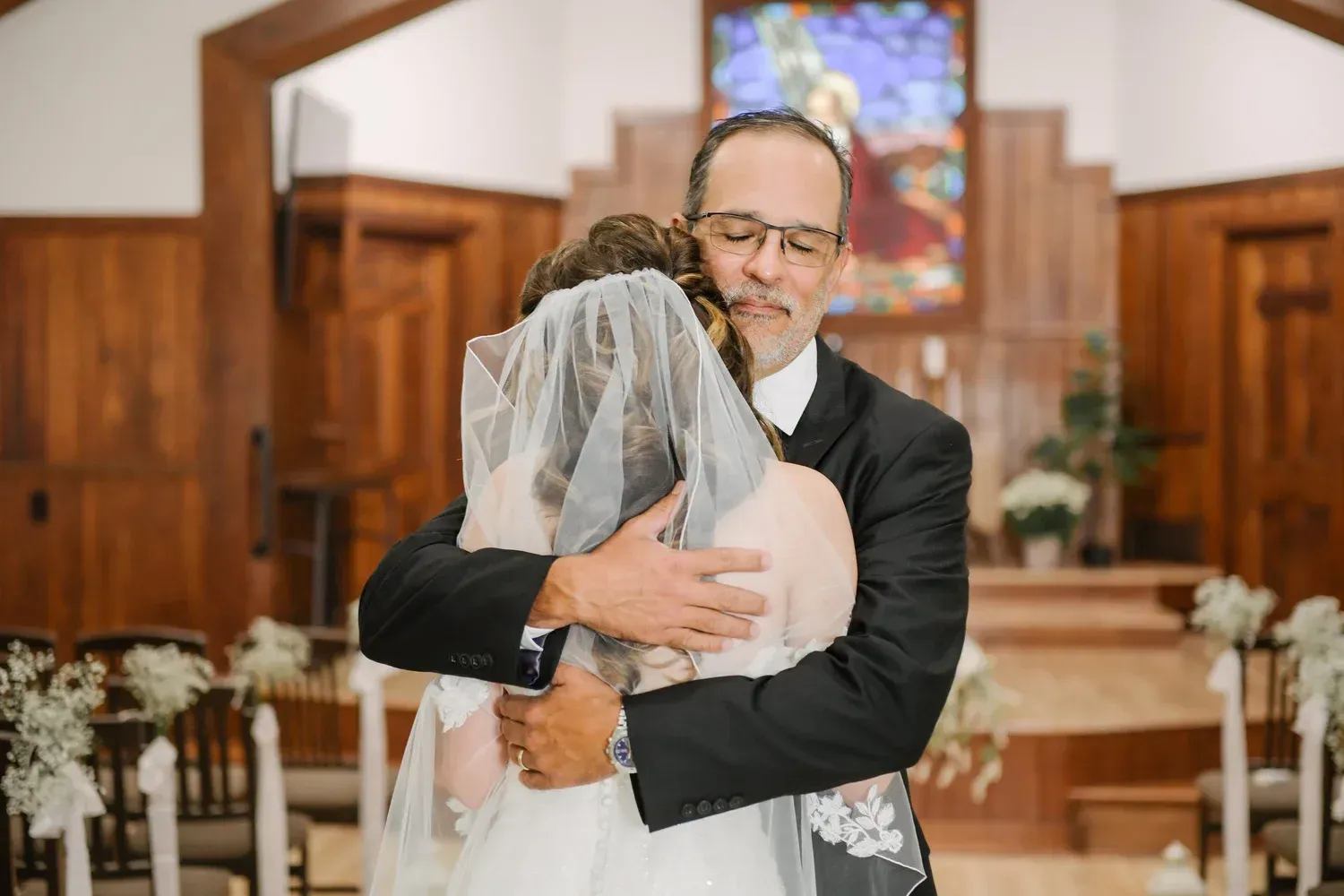 A bride in a white wedding gown and veil hugs a man in a black suit inside a church with wooden paneling.