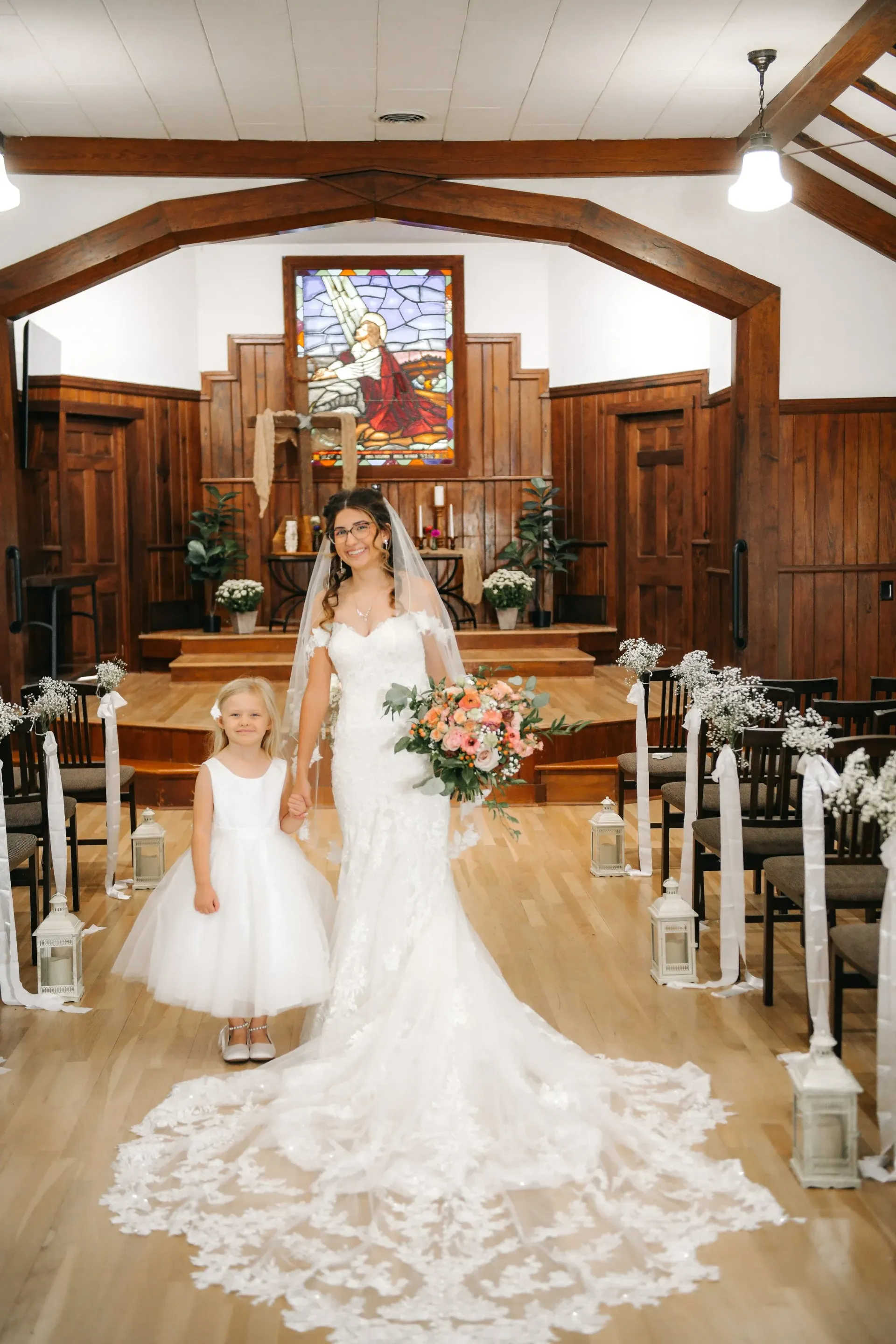 A bride in a lace-trimmed gown and a young attendant stand holding hands in the aisle of a wood-paneled church.