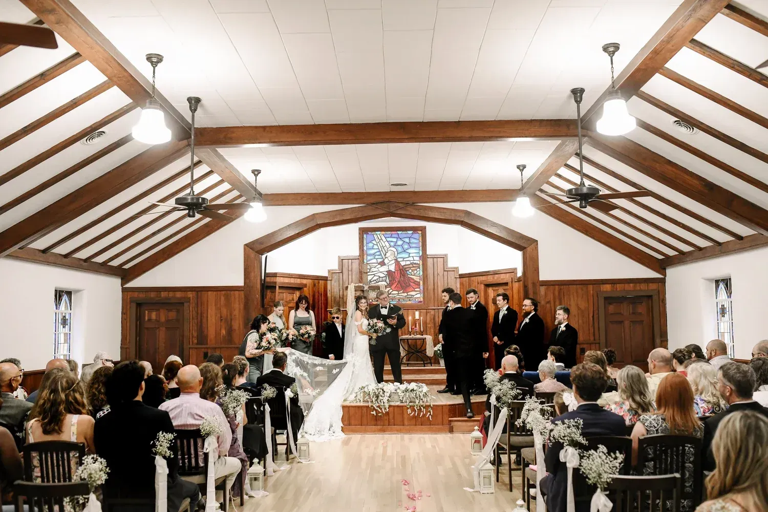 A wedding ceremony takes place in a rustic chapel with wood-paneled walls and exposed beams as guests sit in rows.