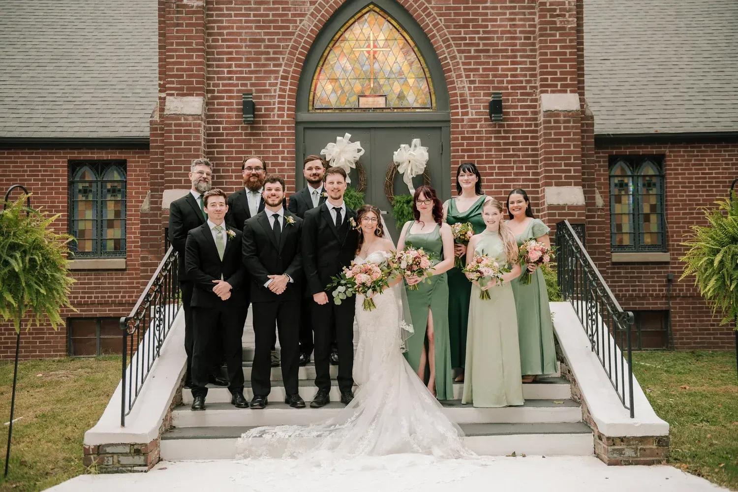 Wedding party posing for a group photo on the steps of a brick church with an arched, stained-glass door.