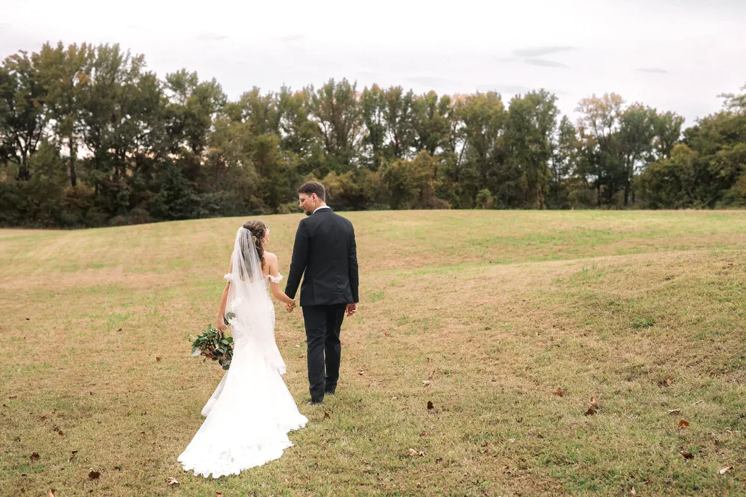 A bride in a white wedding gown and a groom in a black suit walk hand-in-hand across a grassy field toward a line of trees.