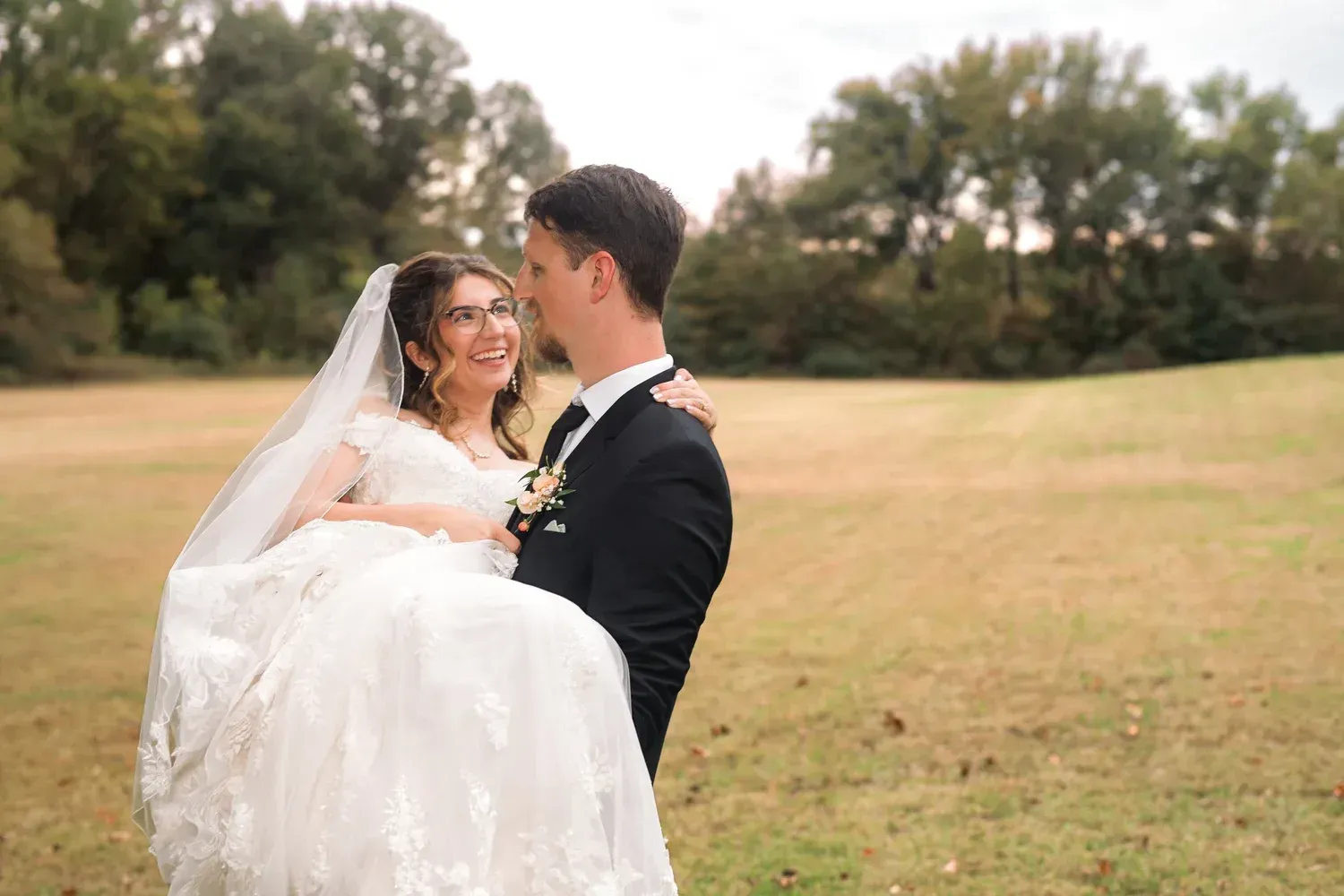 A groom in a black suit carries his bride in a white lace wedding dress through a large, grassy field.