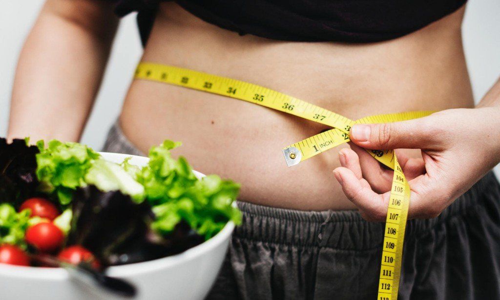 Person measuring waist with yellow tape, holding salad bowl, implying weight loss.