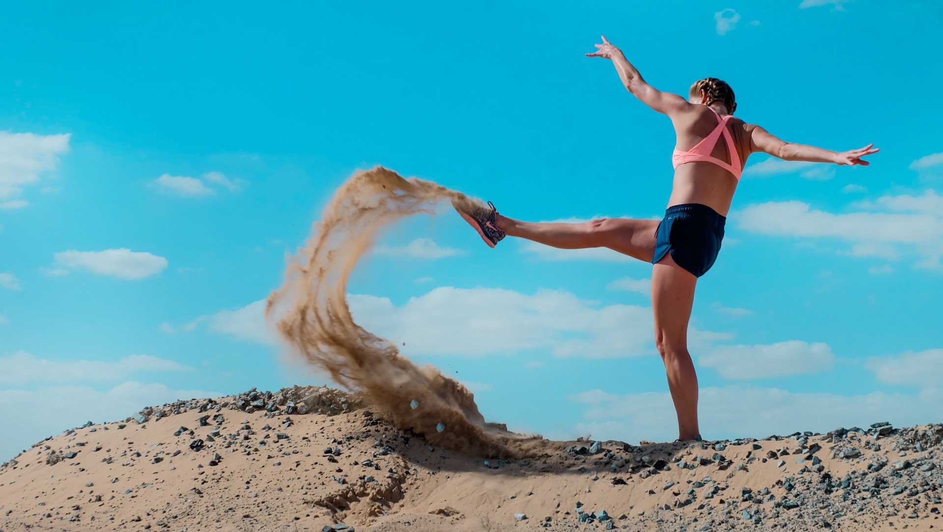 Woman in athletic wear kicking up sand against blue sky.