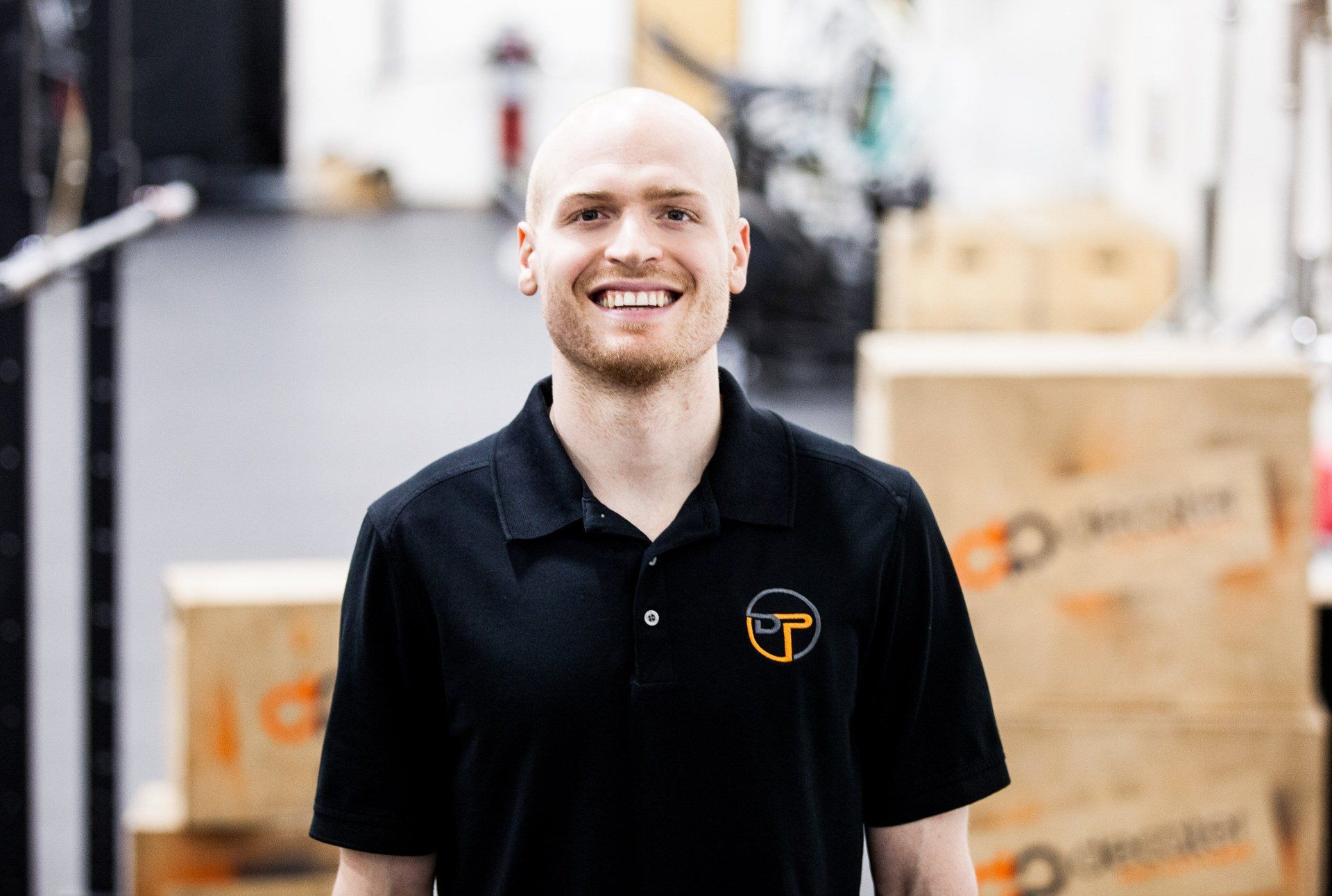 Man with bald head smiling, wearing black polo shirt, standing in gym.