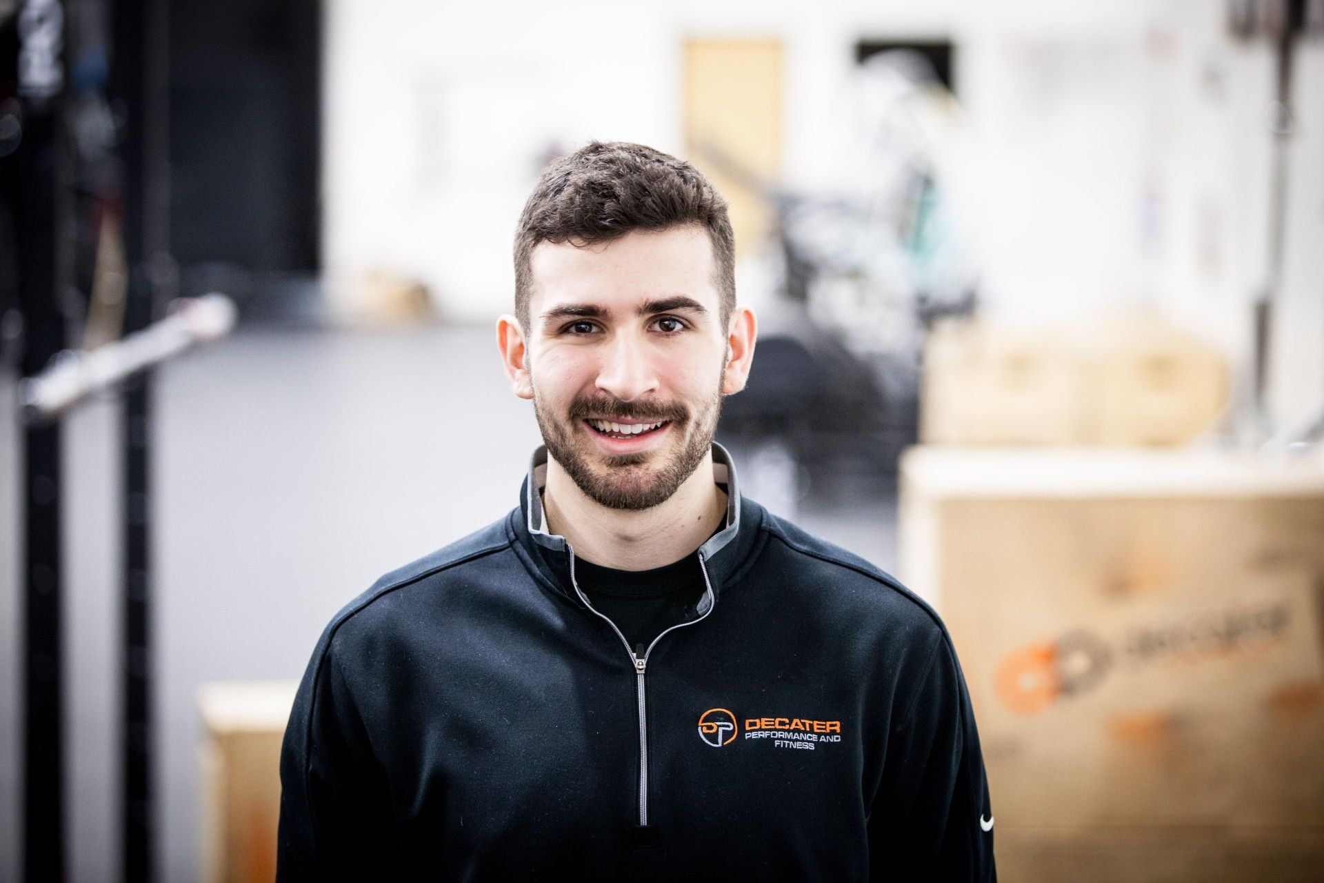 Man with dark hair and beard smiles, wearing a black sweatshirt in a gym setting.