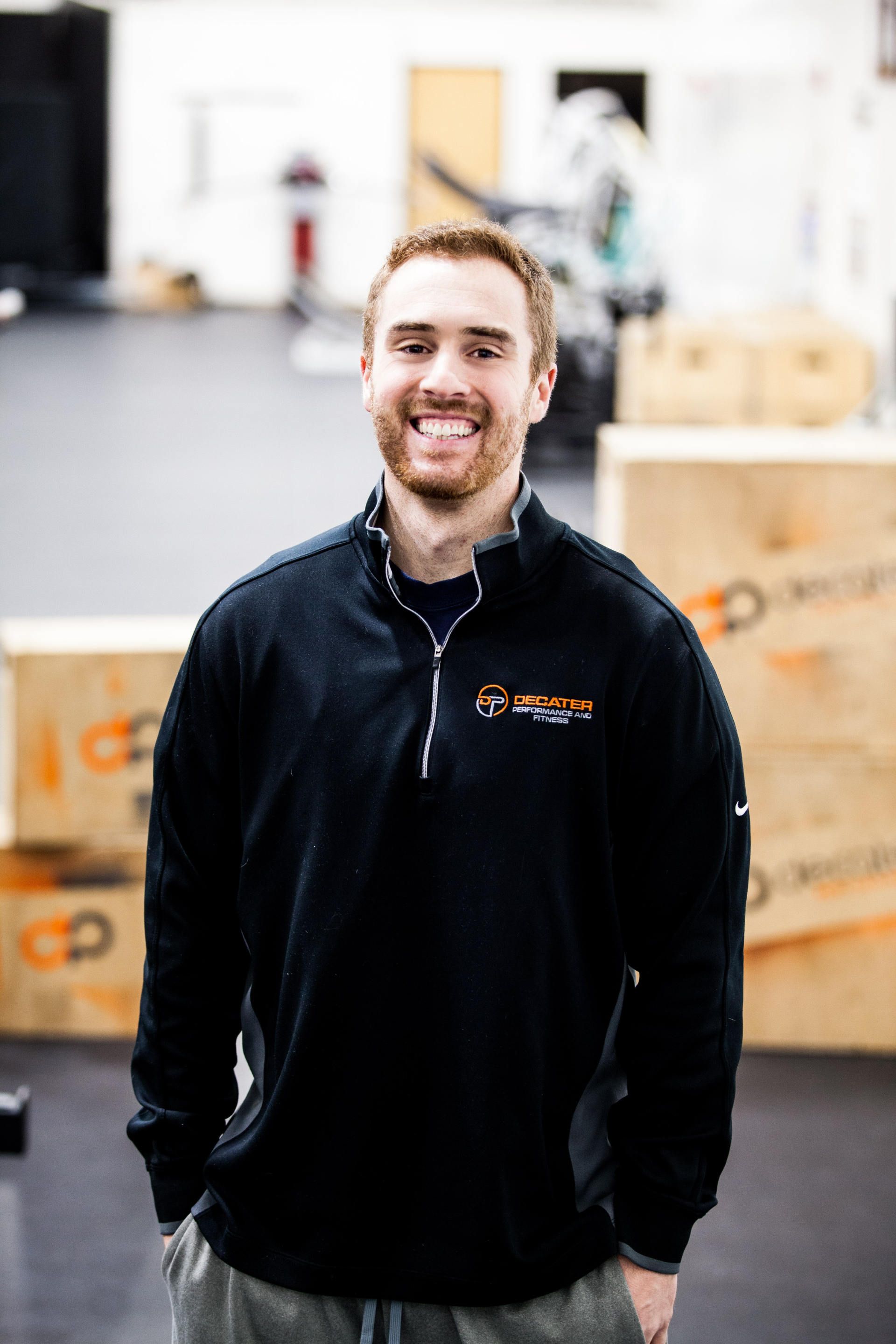 Man with a beard and smile, in black zip-up, stands in a gym, wooden boxes behind him.