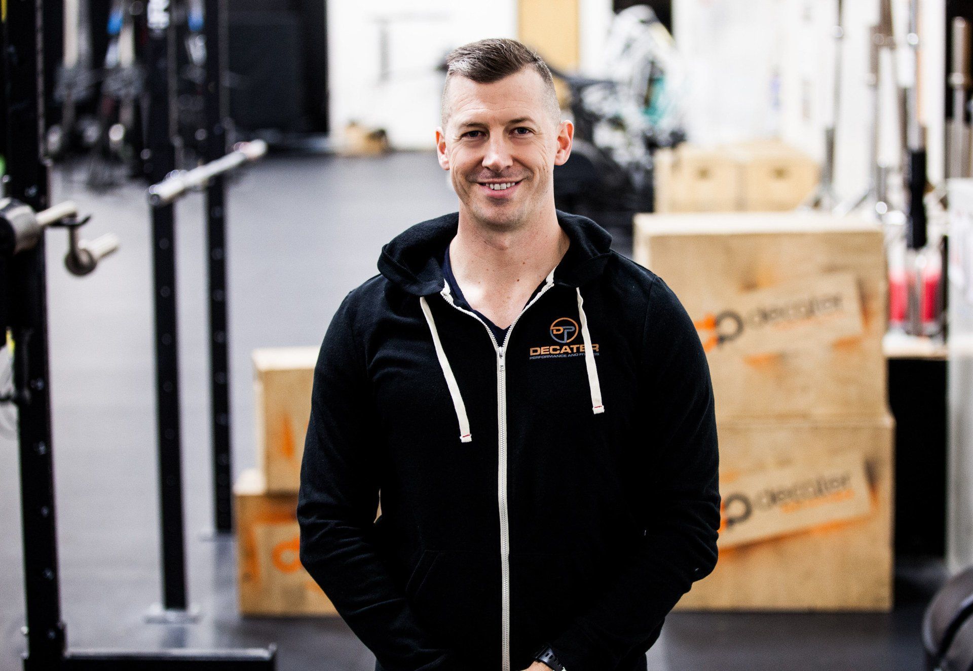 Man in black hoodie smiles in a gym setting, boxes in the background.