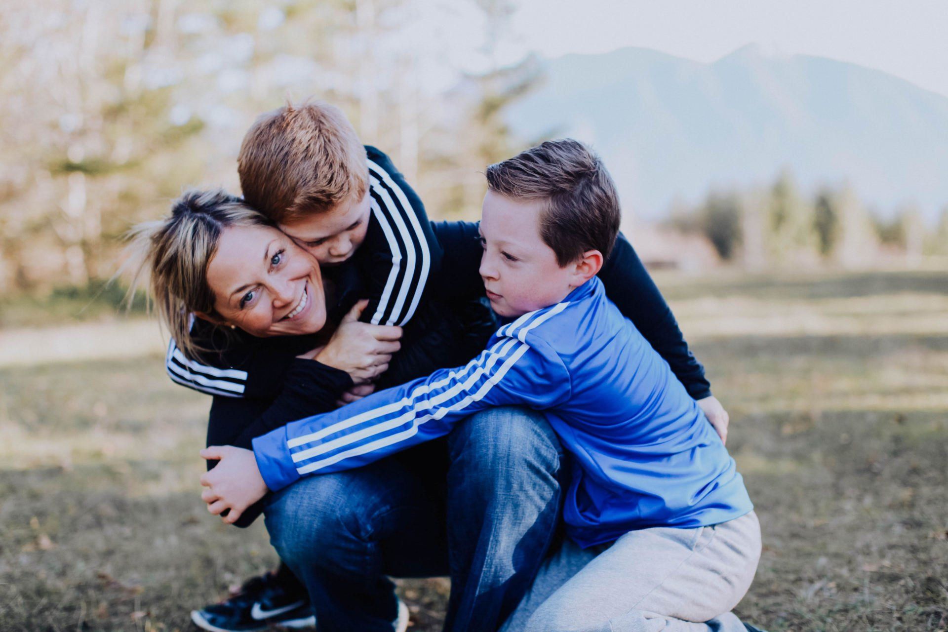 Mother and two sons hugging, smiling outside near mountains. They wear blue, black, and white jackets.