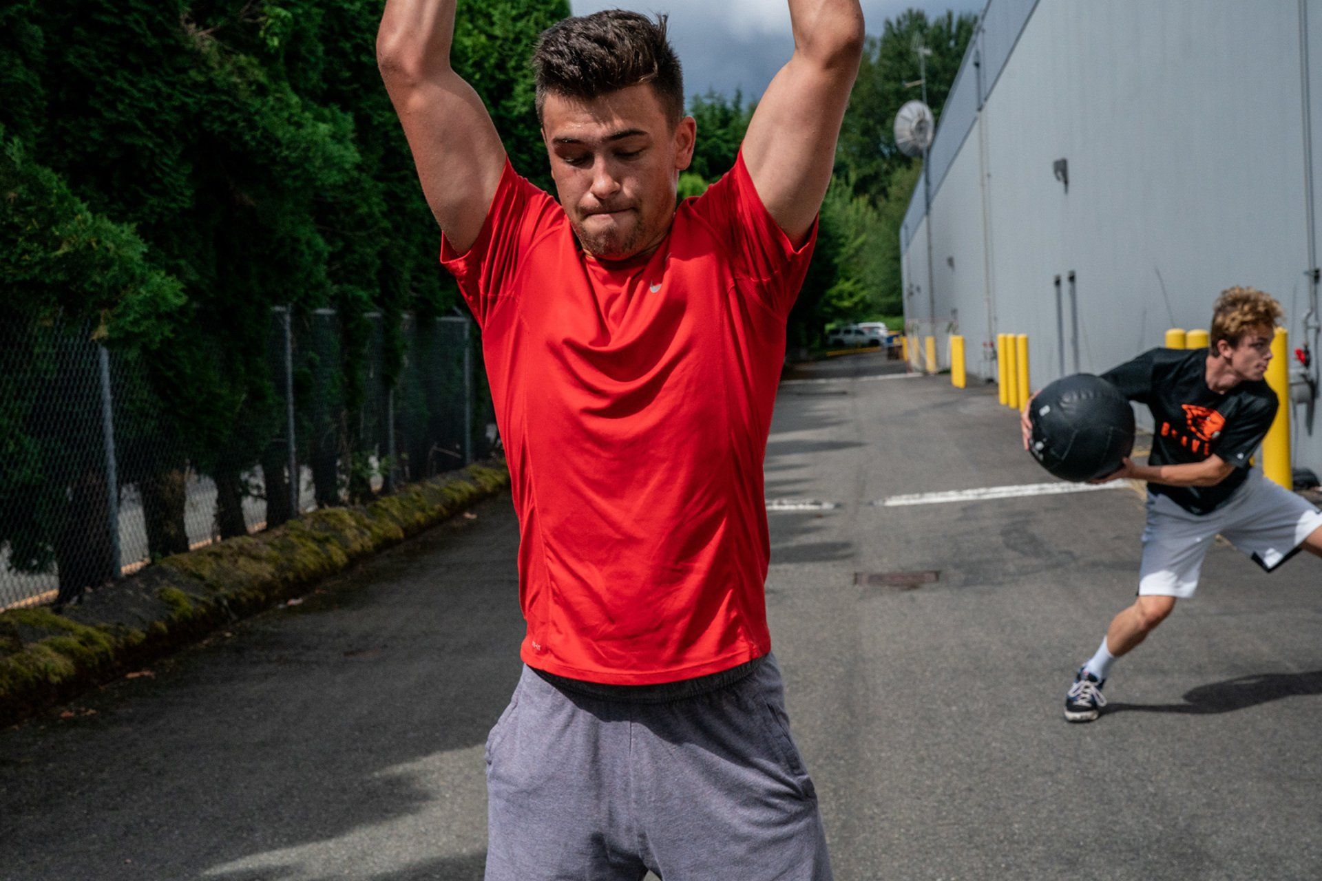 Man in red shirt raising arms, prepares to slam down medicine ball as another man throws it outdoors.