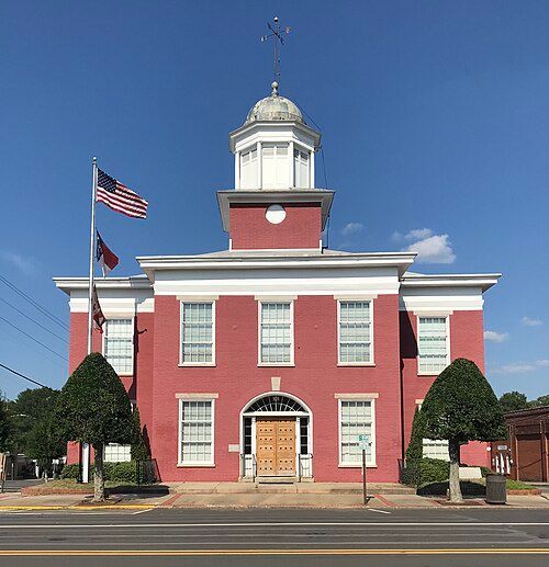 Red brick courthouse with white trim and cupola; American and state flags fly.