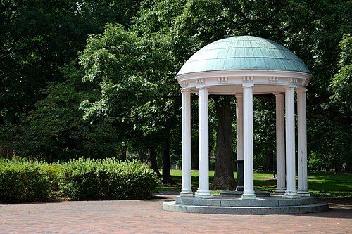 White columns support a teal-roofed, circular gazebo. Green trees surround it on brick ground.