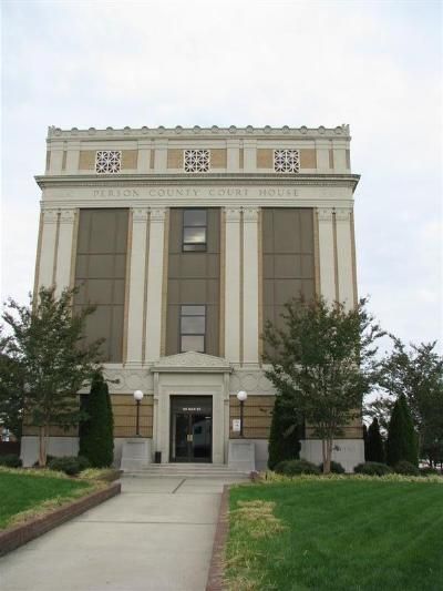 Person County Courthouse, a tall beige building with large windows and a central entrance, framed by trees and a paved path.