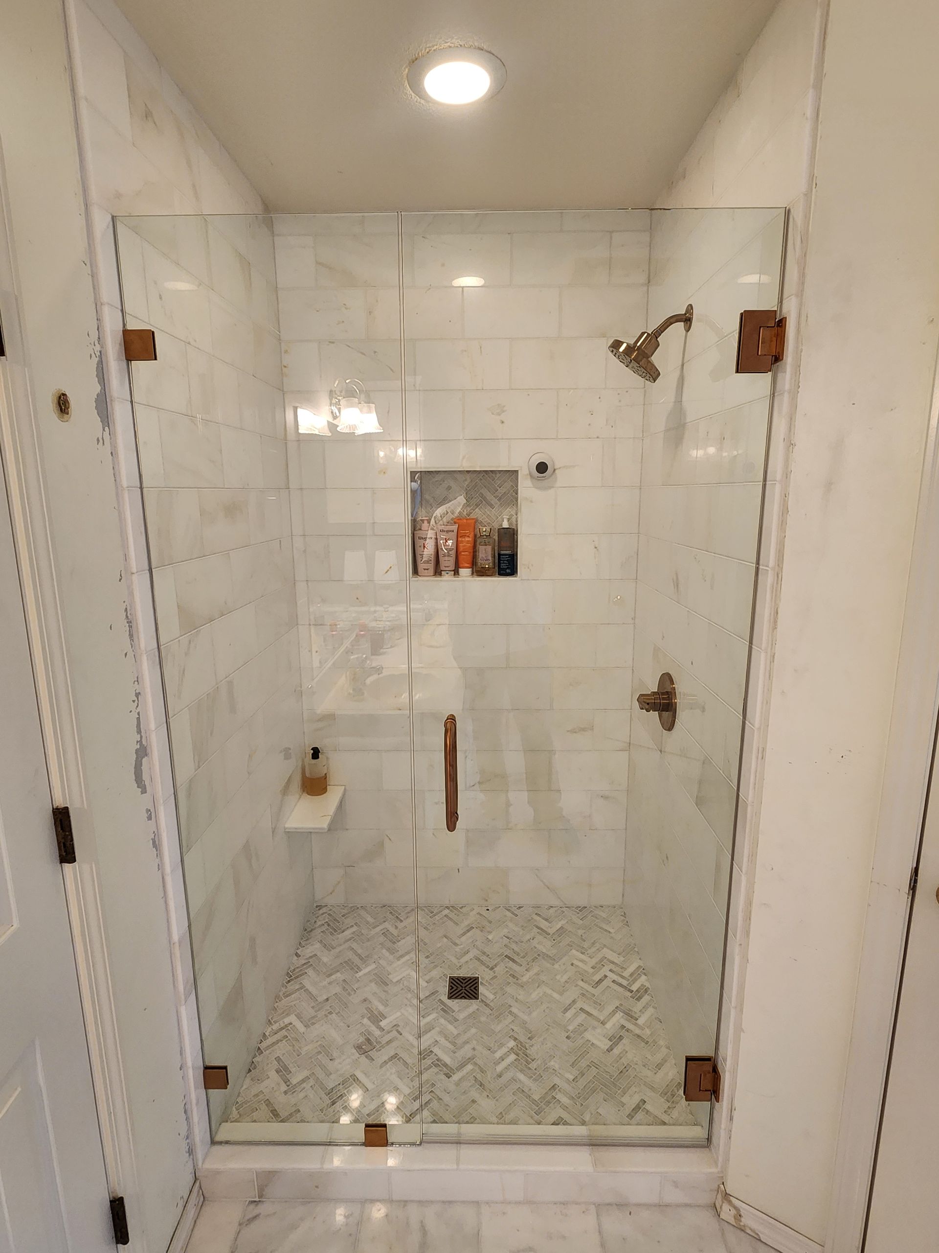 Glass-enclosed shower with white marble tiles and herringbone tile floor, bronze fixtures, and built-in shelf.