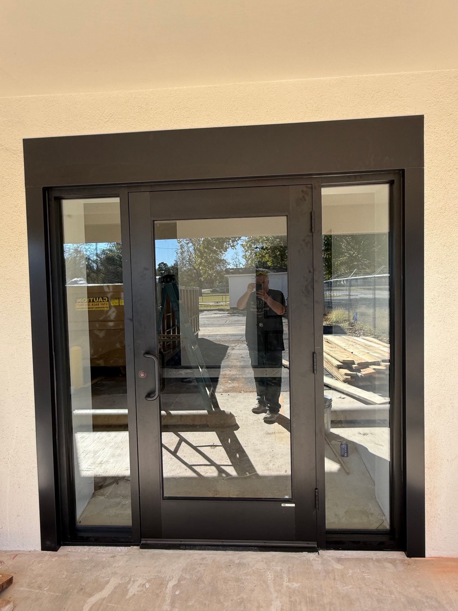 Black framed glass door and sidelights installed in beige exterior wall. Person visible in reflection.