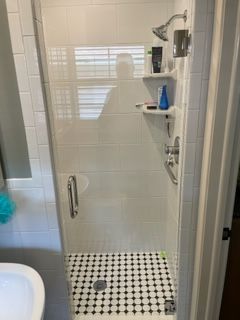 Shower with white tile walls and black and white patterned floor; a glass door, and shelves with toiletries.