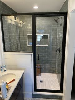 Black-framed shower enclosure with gray tiled walls, white tile floor, and sink in a bathroom.