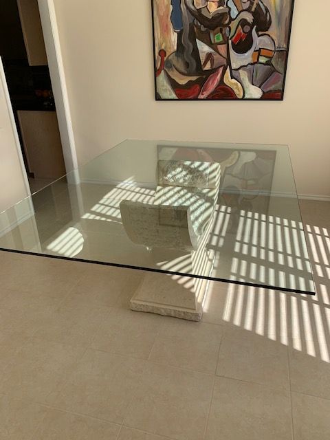 Glass-top dining table on a light-colored stone pedestal, beneath abstract artwork on a beige wall.