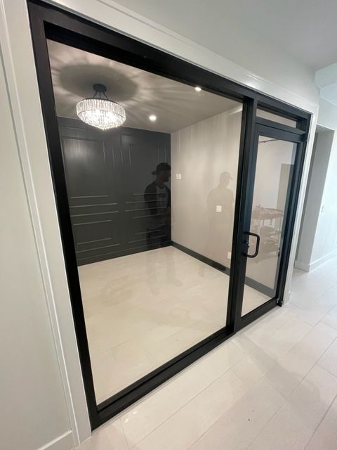 Black-framed glass door and wall, showing a room with a chandelier, gray wall, and white floor; person inside.