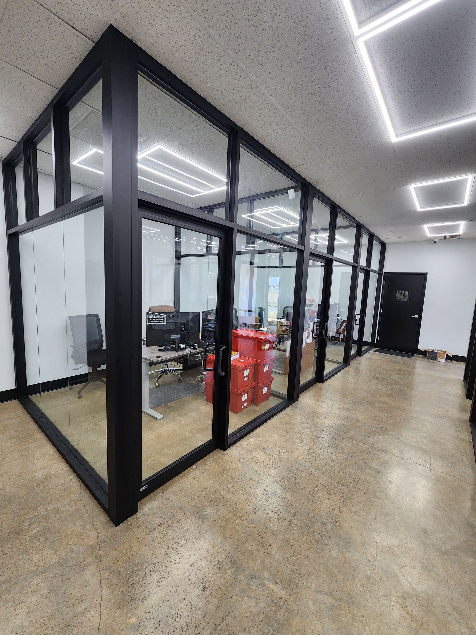Office interior with glass-walled rooms, black frames, and a hallway with a concrete floor.