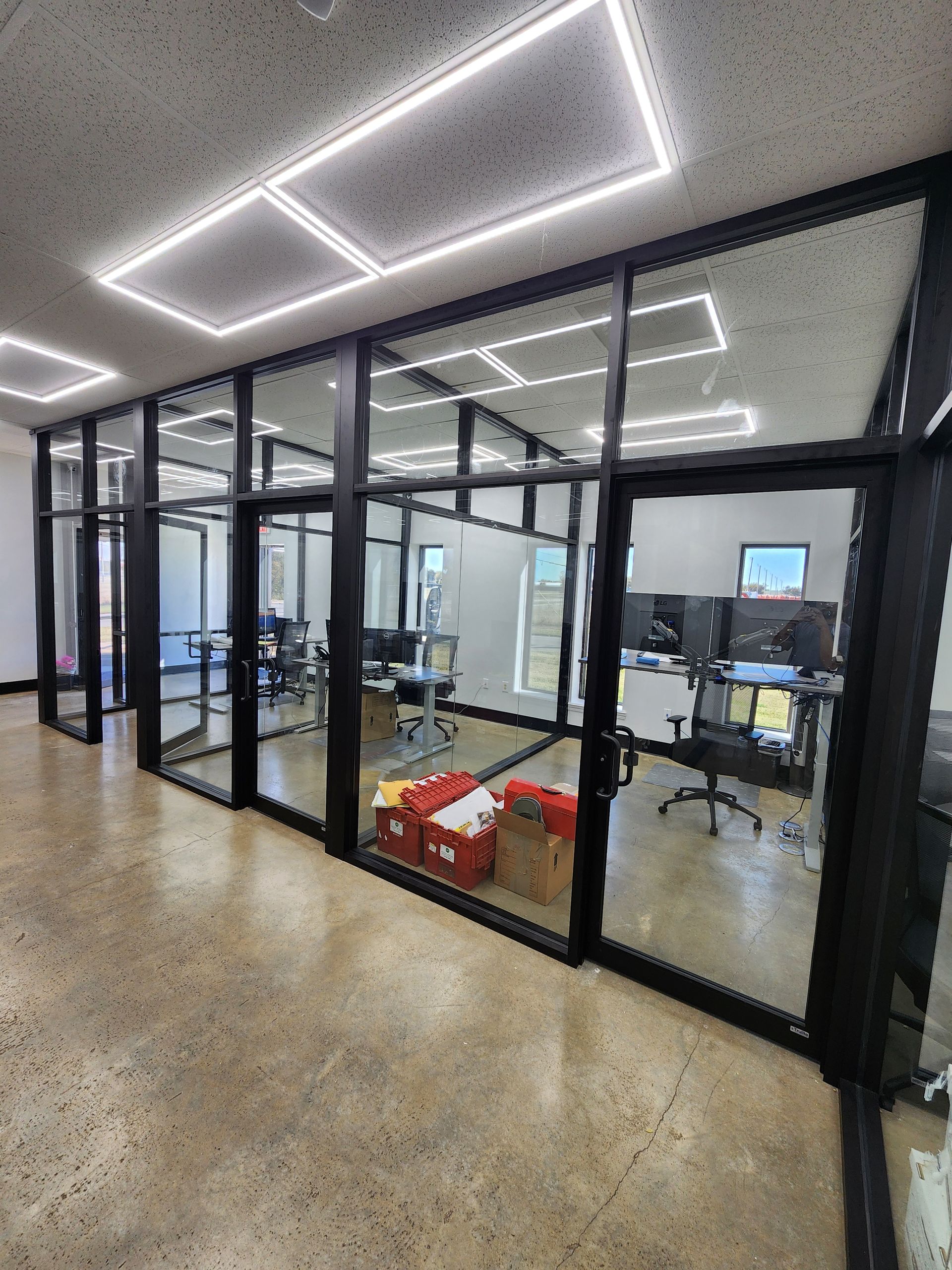 Office space with glass-walled rooms, black frames, and a polished concrete floor. Overhead are rectangular lights.