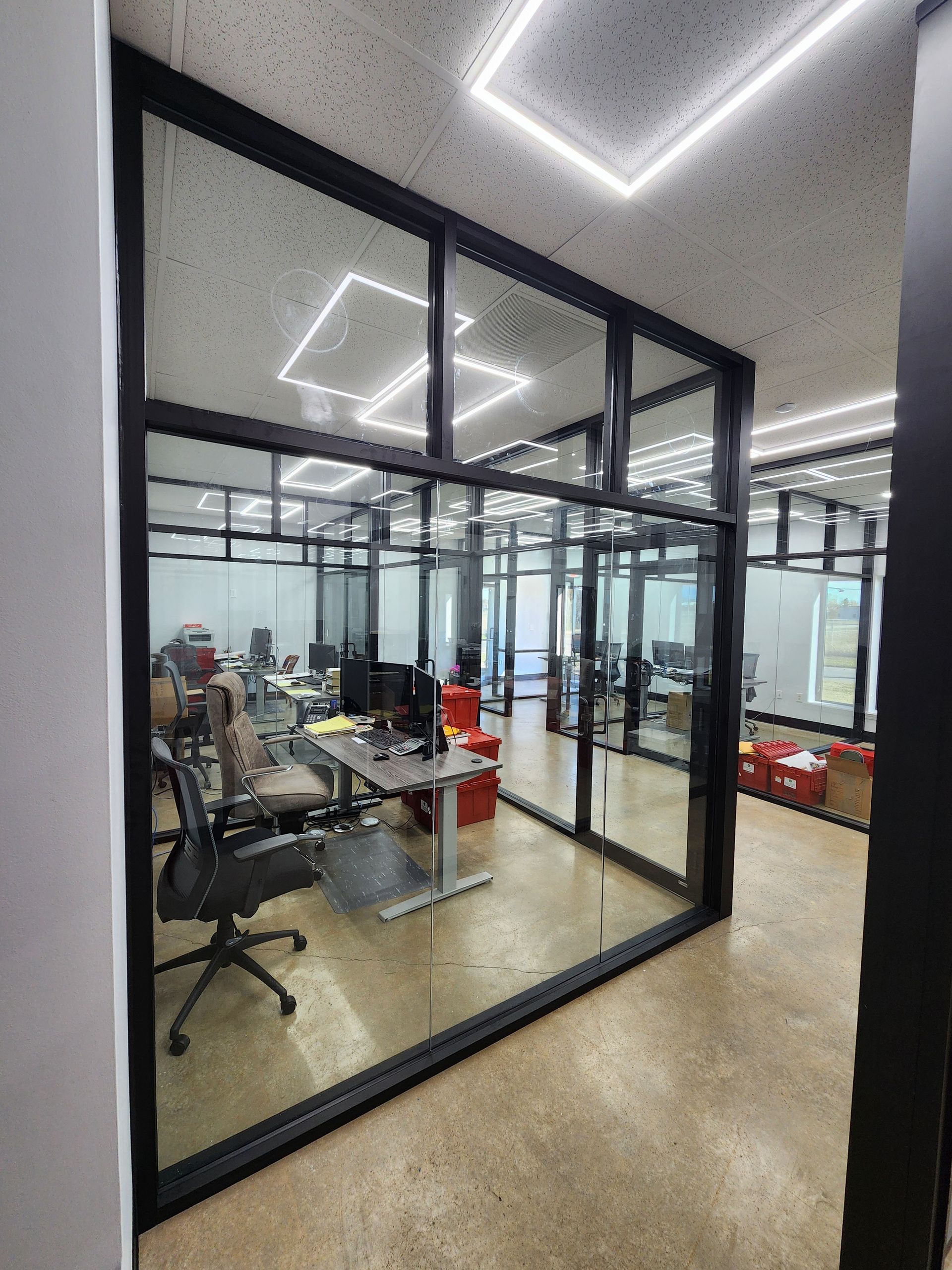 Glass-walled office space with black frames, desks, and chairs visible inside. A hallway leads toward the offices.