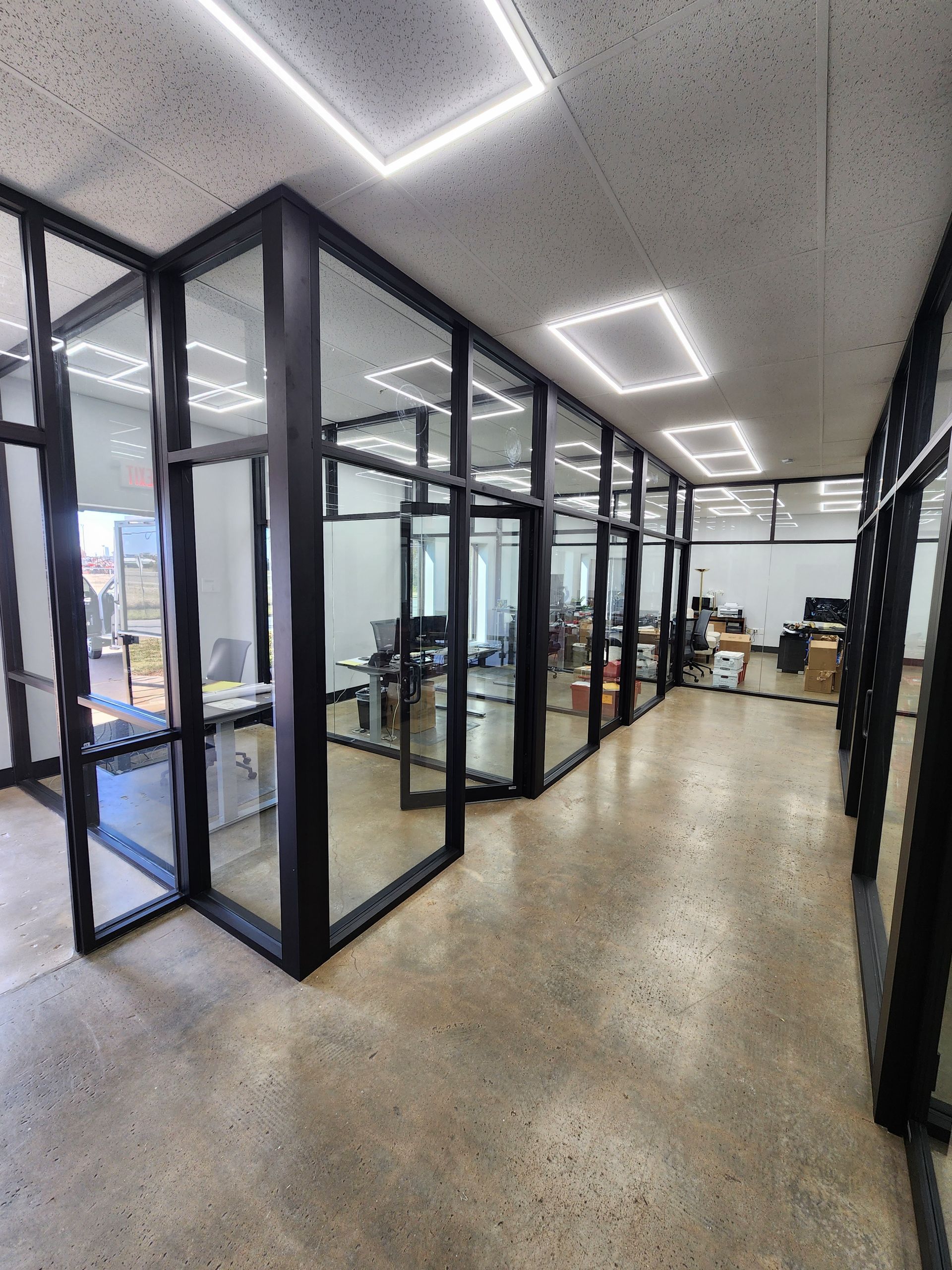 Office hallway with glass-walled offices. Black frames, concrete floor, white ceiling lights.
