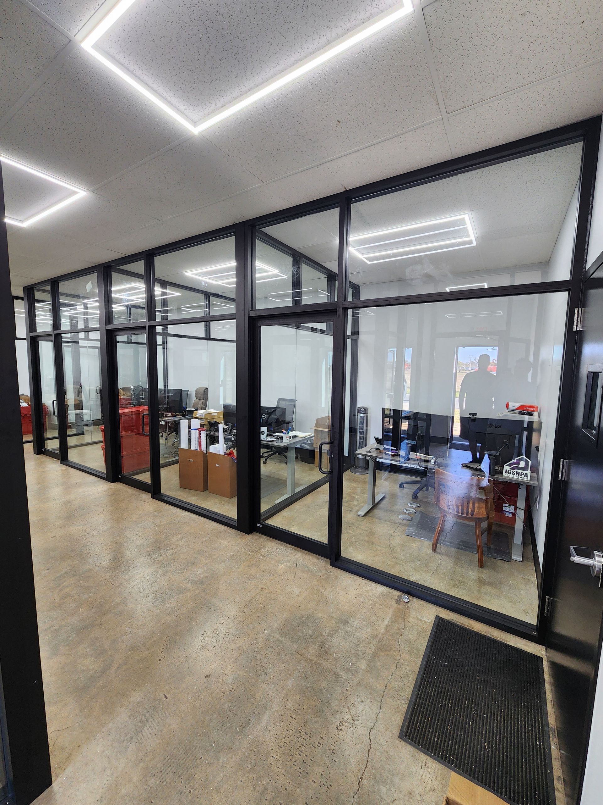 Office with glass-walled rooms, black frames, and a concrete floor. Fluorescent lights and a person visible inside.